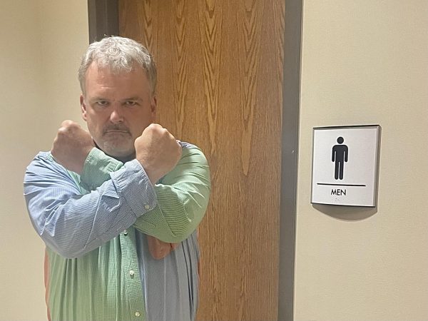Pastor Davis stands in front of the mens bathroom holding his arms in defiance.